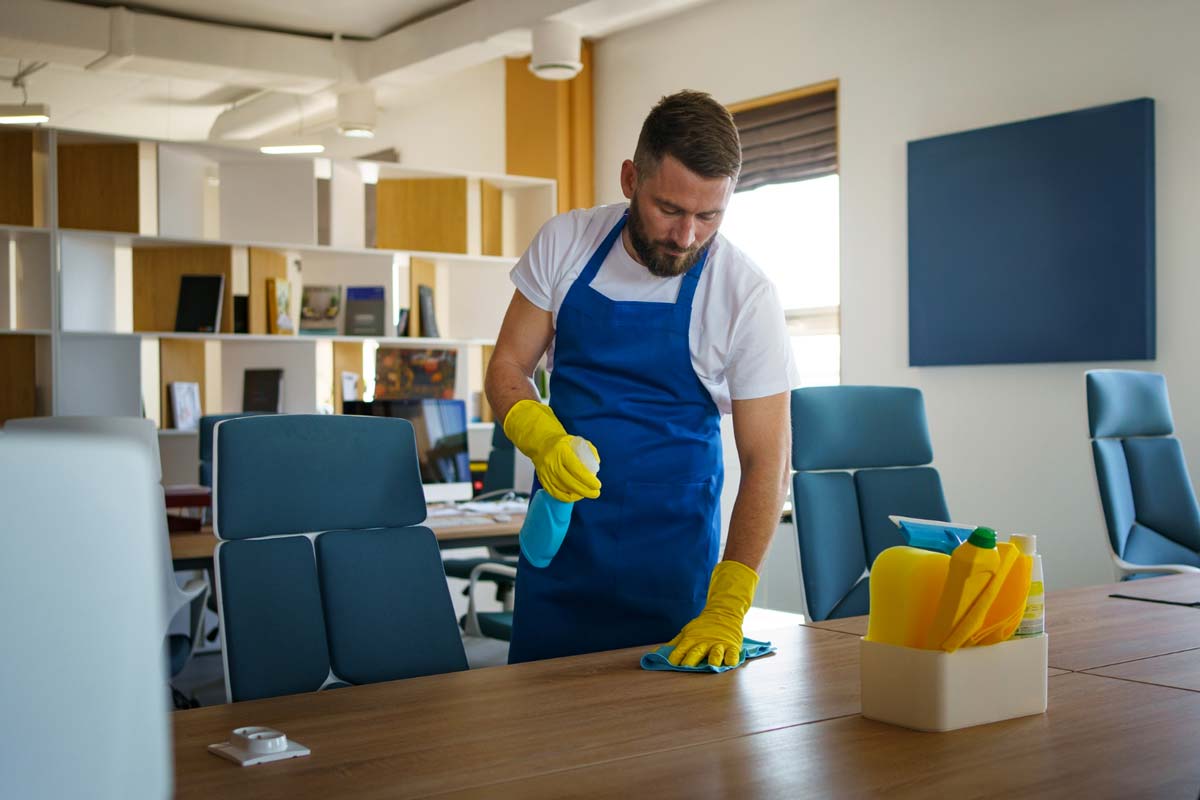A man wearing yellow cleaning gloves and a blue apron wipes a workspace table in an office.